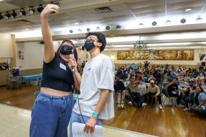 two young people pose for a selfie with a large crowd